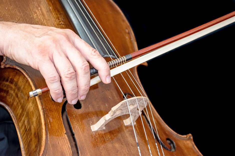 Der Mittelteil eines Cellos vor schwarzem Hintergrund wird von einer männlichen Hand mit Bogen bespielt.