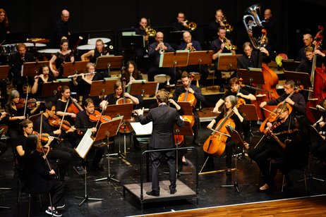 Das Sinfonieorchester sitzt auf der Bühne und spielt unter der Leitung des Dirigenten Marcos Kopf, der vor dem Orchester steht. 