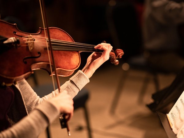 Frauenhände bespielen eine Violine. Rechts im Bild ist ein Teil eines Notenständers zu sehen.