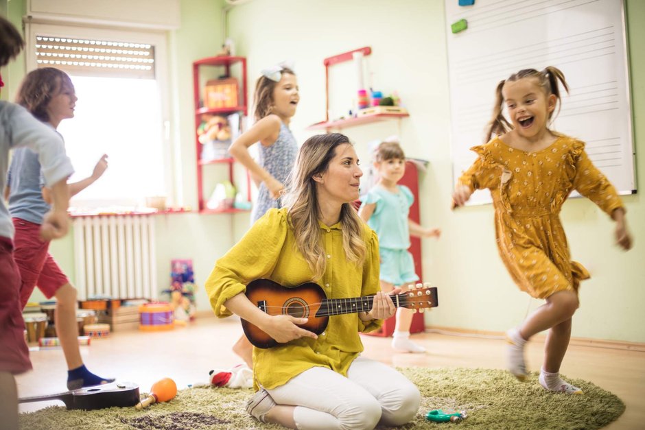 Vier singende Kinder springen um eine Frau herum, die auf einem Teppich kniet und Gitarre spielt