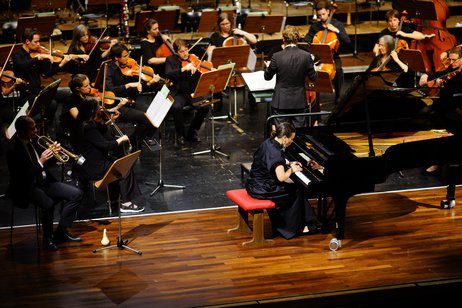Die Solisten Patrick Hofer (Trompete) und Kateryna Titova (Klavier) sitzen auf der Bühne vor dem Sinfonieorchester und spielen auf ihren Instrumenten. 